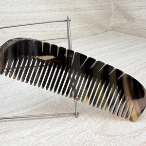 May include: A dark brown comb with a curved handle and closely spaced teeth. The comb is made of a material with a marbled appearance, and it rests on a metal rack. The comb's teeth are evenly spaced and extend from the curved handle.