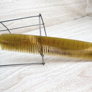 May include: A vintage, olive-toned comb with fine teeth. The comb is made of a translucent material and has a curved shape. It rests on a wire display stand against a light-colored wooden surface.