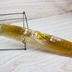 May include: A golden-yellow comb with fine teeth, resting on a light-colored wooden surface. The comb has a smooth, polished appearance and is angled towards the viewer. A dark metal wire structure is partially visible in the background.
