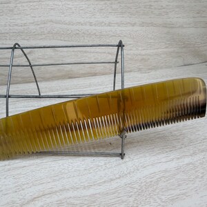 May include: A golden-yellow comb with fine teeth, transitioning to a darker shade at the end. The comb is displayed on a metal wire rack against a light wood-grain background. This hair care accessory is designed for grooming.