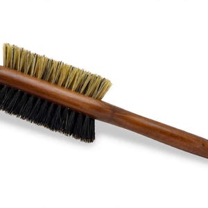 May include: A double-sided brush with a polished wooden handle. One side features stiff, light beige bristles, while the opposite side features dense, black bristles. The brush has a long, slender handle and a rectangular head design.