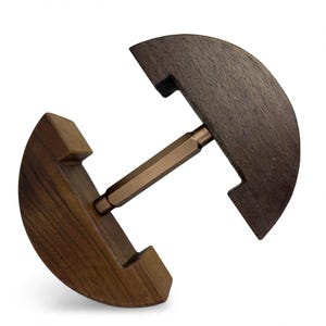 May include: A dumbbell-shaped object consisting of two dark brown, semi-circular wooden blocks connected by a central, hexagonal, metallic bronze-colored rod. The wooden pieces feature a flat inner edge with a rectangular notch cut out.