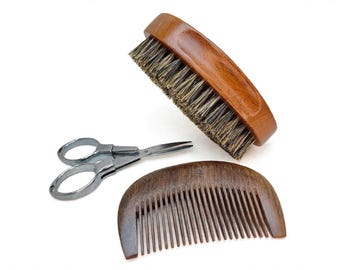 Può includere: Un set per la cura personale composto da una spazzola ovale in legno lucido con setole scure, un pettine in legno marrone scuro a denti fini e un paio di forbici in metallo argentato con anelli arrotondati, disposti su sfondo bianco.