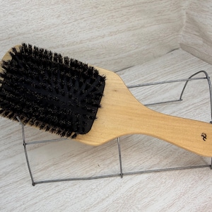 May include: A wooden hair brush with a light-colored handle and a rectangular head filled with black bristles. The brush is resting on a metal wire display stand. The handle has a small logo.