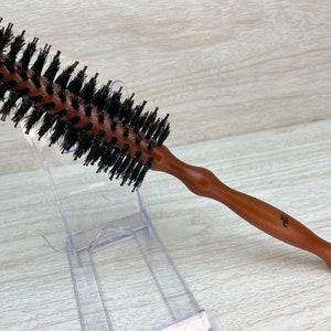 May include: A round hair brush with a wooden handle and black bristles. The brush is designed for styling and grooming hair. The handle is a medium brown color and has a curved design for easy grip.