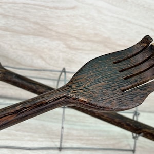 May include: Close-up of two wooden kitchen utensils. One is a spoon, and the other is a fork-like utensil with a hand-shaped design. Both are dark brown with visible wood grain. The utensils are resting on a wire rack.