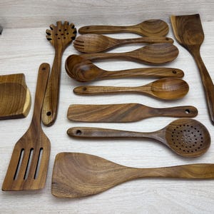May include: A collection of wooden kitchen utensils, including spoons, spatulas, and a pasta fork. The utensils are made of a light brown wood and are arranged on a light-colored surface. The set includes a spoon rest.