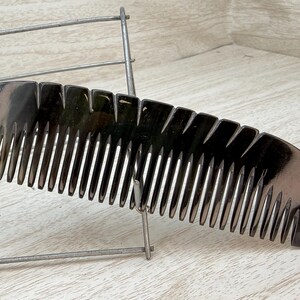 May include: A dark, glossy comb with a curved handle and closely spaced teeth. The comb is black with a hint of yellow at the base. The teeth are evenly spaced and the comb is resting on a metal rack.