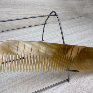 May include: A light brown comb with a curved handle and fine teeth. The comb is made of a translucent material with vertical grooves on the handle. The comb is resting on a metal wire rack.