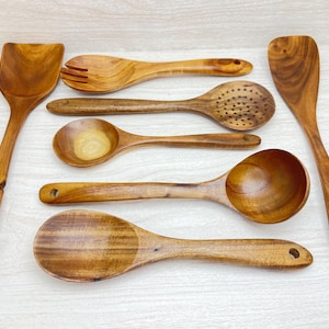 May include: A collection of wooden kitchen utensils, including a spatula, slotted spoon, fork, and various spoons. The utensils are made of a light brown wood and have a smooth finish. Each utensil has a hole in the handle for hanging.