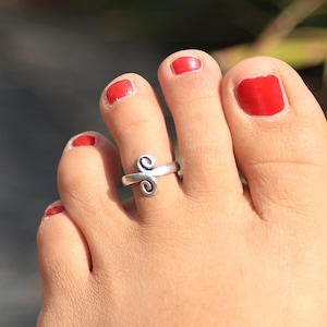 May include: A silver toe ring with a swirl design. The ring is worn on the second toe of a foot with red nail polish.