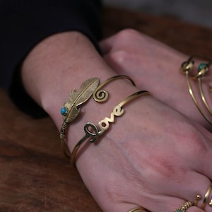 Può includere: Un set di braccialetti dorati con un ciondolo a forma di piuma e una pietra turchese. Il braccialetto presenta anche la parola "love" incisa su di esso.