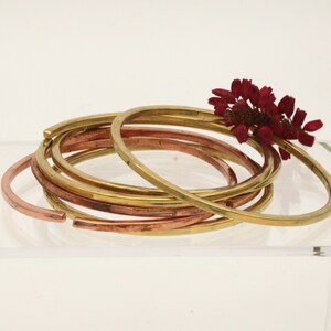 May include: A set of six gold and copper bangles stacked together. The bangles are smooth and shiny, and they are arranged in a loose pile. A small cluster of dried red flowers is resting on top of the bangles.