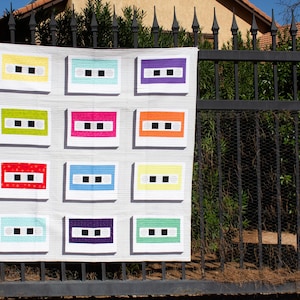 Puede incluir: Una colcha blanca con un patrón repetido de cintas de casete. Cada cinta tiene un fondo de color diferente, incluyendo amarillo, verde, rojo, azul, naranja, morado y verde claro.
