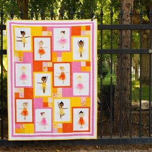 May include: A pink and yellow quilt with a patchwork design featuring multiple images of ballerinas in various poses. The ballerinas are all wearing pink tutus and have brown skin.