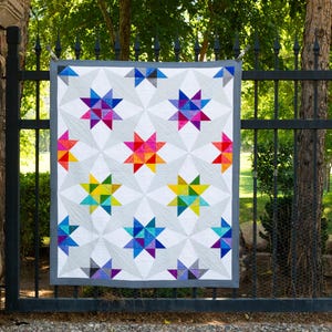 May include: A colourful quilt with a geometric star pattern in shades of blue, green, yellow, orange, red, and purple. The quilt is hanging on a black metal fence.