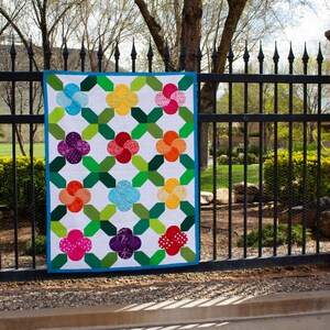 May include: A colorful quilt with a floral pattern hangs on a black metal fence. The quilt features a white background with green cross shapes and flower-shaped designs in various colors, including pink, purple, orange, yellow, and blue. A blue border frames the quilt.