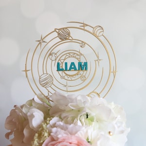 May include: Gold cake topper with a solar system design and the name "LIAM" in teal blue letters. The topper is on top of a white cake with white and pink flowers.