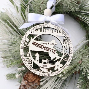 May include: A white wooden Christmas ornament shaped like the state of Massachusetts with the text "MASSACHUSETTS" and "Henry & Lucy" inside the state outline. The ornament also features a lighthouse, a house, and other landmarks around the state. The text "2021" is at the bottom of the ornament. The ornament is hanging on a white ribbon.