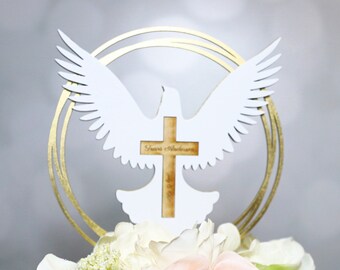 May include: White dove with outstretched wings holding a wooden cross with the text "Jesus is the Answer" in gold lettering. The dove is perched on a gold circular stand with a floral arrangement of white and pink flowers.