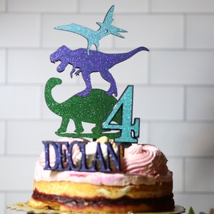 May include: A pink and white frosted cake with a green, purple, and blue glitter dinosaur topper. The topper includes a dinosaur, a pterodactyl, and the number 4. The cake is decorated with the name "Declan".