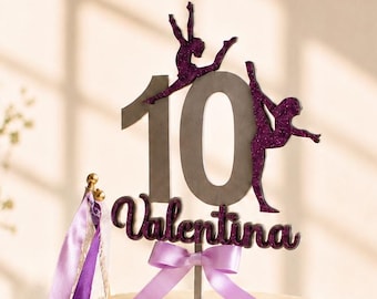May include: A cylindrical cake with textured white frosting features a custom topper displaying the number 10, the name Valentina in purple glitter, and two gymnastic silhouettes. A light purple ribbon bow and a rhythmic gymnastics ribbon are attached.