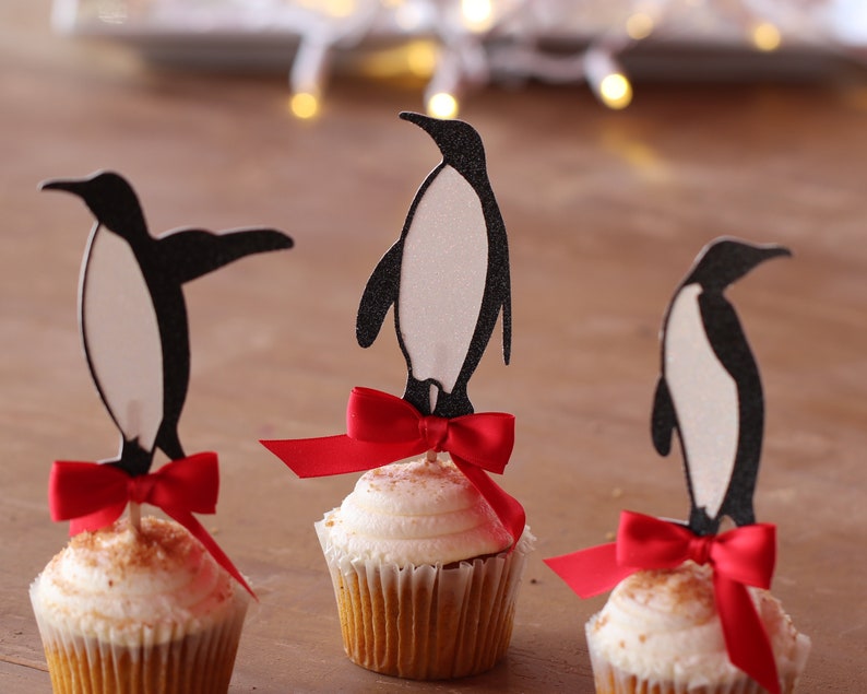 Pu&ograve; includere: Tre cupcake decorati con topper a forma di pinguino nero e fiocchi rossi. I cupcake hanno glassa bianca e granella di zucchero marrone.