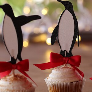 Pu&ograve; includere: Tre cupcake decorati con topper a forma di pinguino e fiocchi rossi. I pinguini sono neri e bianchi e i cupcake sono glassati con glassa bianca e cosparsi di briciole marroni.