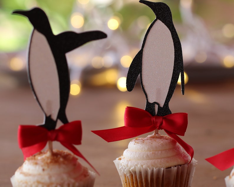 Pu&ograve; includere: Tre cupcake decorati con topper a forma di pinguino e fiocchi rossi. I pinguini sono neri e bianchi e i cupcake sono glassati con glassa bianca e cosparsi di briciole marroni.