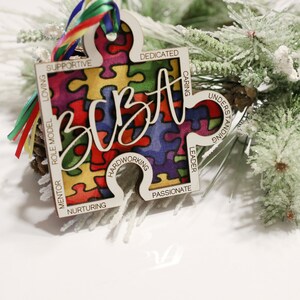 May include: A wooden puzzle piece ornament with a colourful puzzle pattern and the word "BEST" written in cursive. The ornament has words around the edge, including "supportive", "loving", "role model", "mentor", "nurturing", "hardworking", "passionate", "leader", "caring", "understanding", and "dedicated".