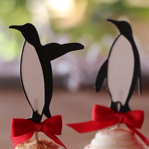 Pu&ograve; includere: Due topper per cupcake a forma di pinguino bianco e nero con fiocchi rossi. I pinguini sono in piedi su cupcake con glassa bianca.