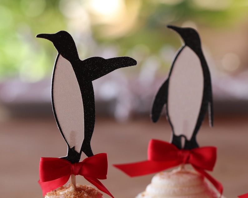 Pu&ograve; includere: Due topper per cupcake a forma di pinguino bianco e nero con fiocchi rossi. I pinguini sono in piedi su cupcake con glassa bianca.