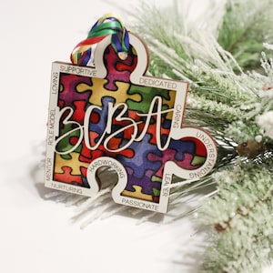 May include: A wooden puzzle piece ornament with a colourful puzzle pattern and the word "best" written in white. The ornament has words around the edge, including "supportive", "loving", "role model", "mentor", "nurturing", "hardworking", "passionate", "leader", "caring", "understanding", and "dedicated".