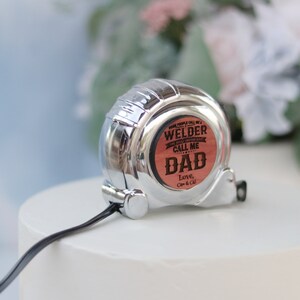 May include: A silver retractable tape measure with a wooden insert that reads "Some people call me a welder, the most important call me Dad. Love, Cap & Cal."
