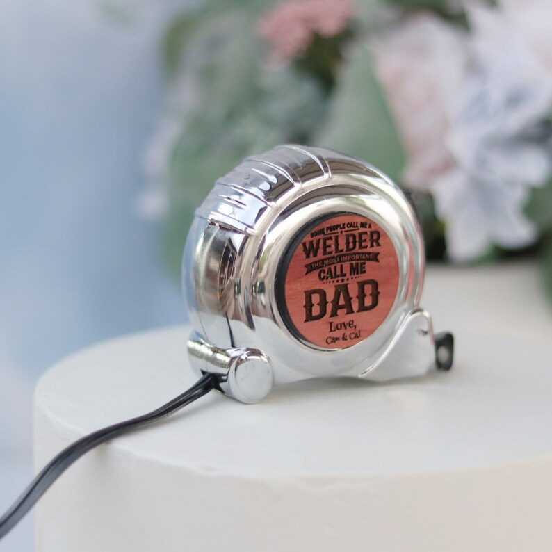 May include: A silver retractable tape measure with a wooden insert that reads "Some people call me a welder, the most important call me Dad. Love, Cap & Cal."