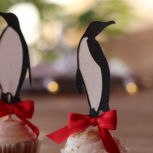 Pu&ograve; includere: Due cupcake sormontati da silhouette di pinguini neri e fiocchi rossi. I cupcake sono glassati con glassa bianca e hanno una base marrone.