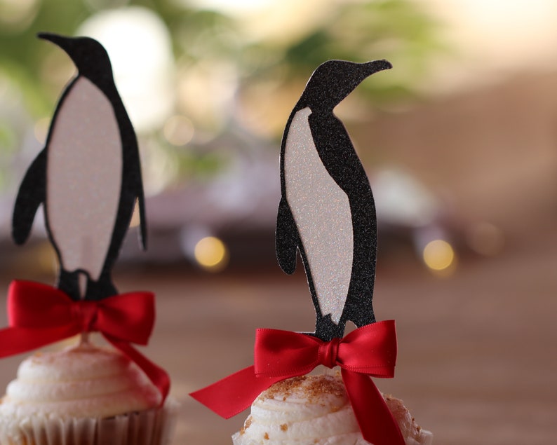 Pu&ograve; includere: Due cupcake sormontati da silhouette di pinguini neri e fiocchi rossi. I cupcake sono glassati con glassa bianca e hanno una base marrone.