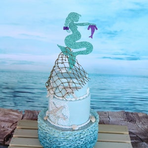 May include: A two-tiered cake decorated with a blue and white seashell design, a brown fishing net, and a glitter mermaid topper holding a small mermaid. The cake is on a wooden crate.
