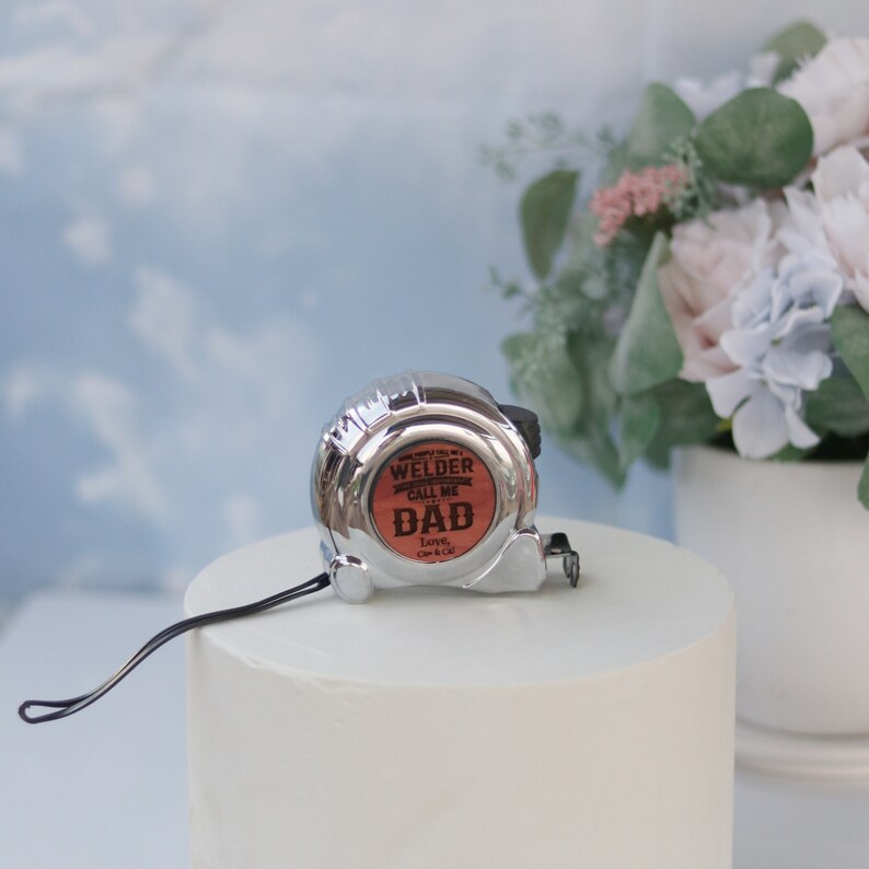 May include: A silver tape measure with a red and black label that reads "Welder Call Me Dad Love, Mom & Dad".