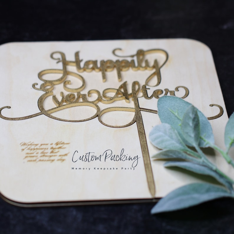 Peut inclure: Un g&acirc;teau topper en bois clair avec le texte dor&eacute; Happily Ever After en &eacute;criture cursive. La base comporte le texte imprim&eacute; Custom Packing et Memory Keepsake Party, accompagn&eacute; d'un petit brin de feuilles vertes sur le c&ocirc;t&eacute;.