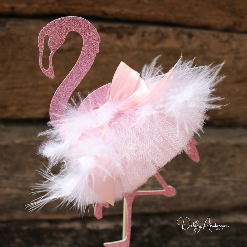 K&ouml;nnte beinhalten: Rosa glitzernder Flamingo-Kuchentopper mit einem wei&szlig;en Feder- und rosa T&uuml;llbogen. Der Topper befindet sich auf einem Holzgrund.
