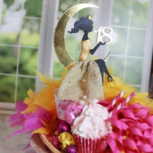 Peut inclure: Une d&eacute;coration de g&acirc;teau repr&eacute;sentant la silhouette noire d'une figure couronn&eacute;e assise sur un croissant de lune dor&eacute; paillet&eacute;. La figure tient le chiffre 18 en blanc. L'ensemble est orn&eacute; de tissu dor&eacute;, de plumes jaunes et roses et de rubans.