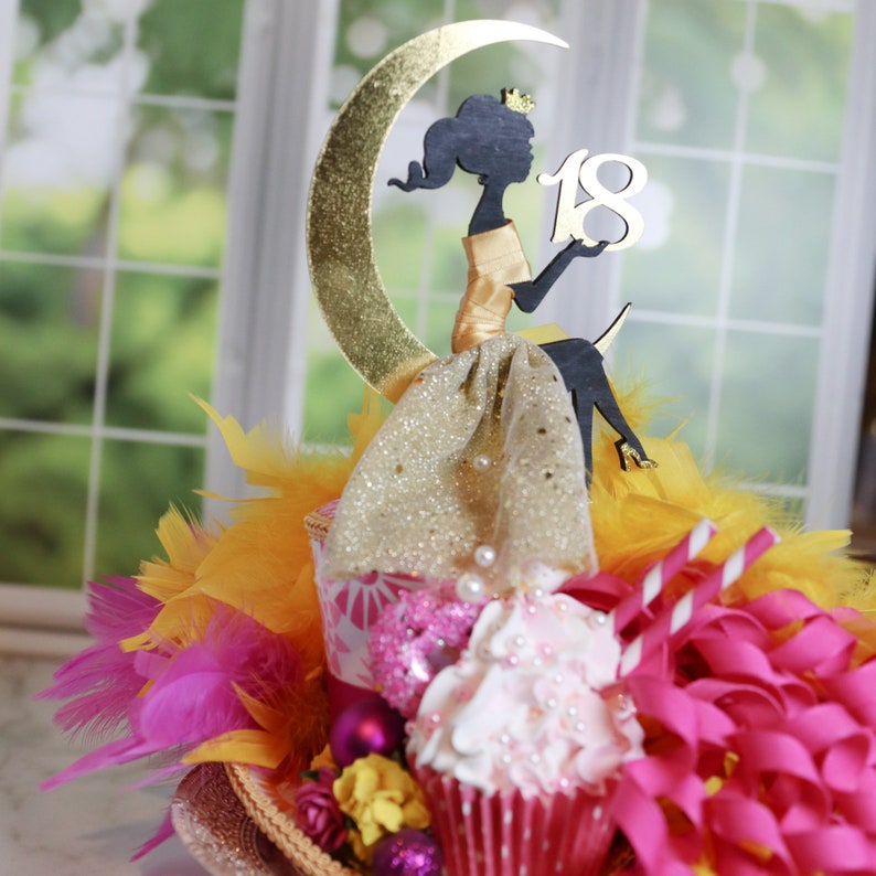 Peut inclure: Une d&eacute;coration de g&acirc;teau repr&eacute;sentant la silhouette noire d'une figure couronn&eacute;e assise sur un croissant de lune dor&eacute; paillet&eacute;. La figure tient le chiffre 18 en blanc. L'ensemble est orn&eacute; de tissu dor&eacute;, de plumes jaunes et roses et de rubans.
