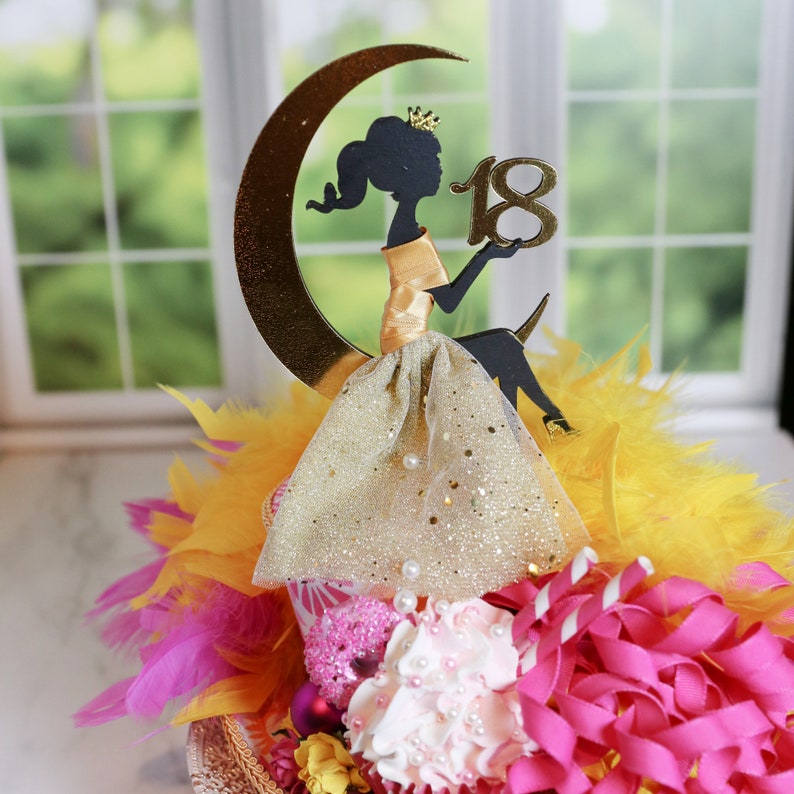Peut inclure: Une d&eacute;coration de g&acirc;teau repr&eacute;sentant la silhouette noire d'une personne couronn&eacute;e assise sur un croissant de lune dor&eacute;, tenant le nombre 18 en or. La figurine porte une jupe en tulle dor&eacute; paillet&eacute; orn&eacute;e de petites perles blanches sur un socle.