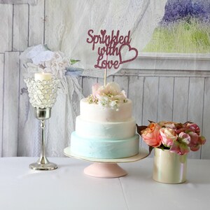 May include: A three-tiered wedding cake with a light blue and white ombre frosting. The cake is decorated with fresh pink and white flowers and a topper that says "Sprinkled with Love".