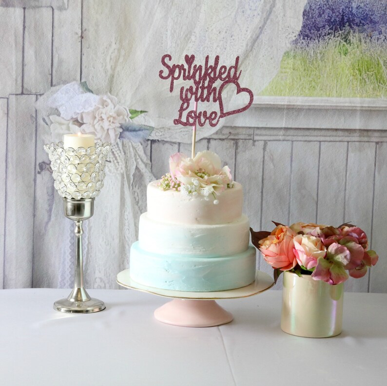 May include: A three-tiered wedding cake with a light blue and white ombre frosting. The cake is decorated with fresh pink and white flowers and a topper that says "Sprinkled with Love".