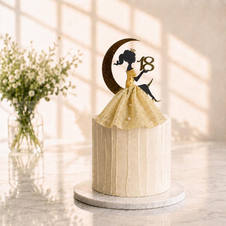 Peut inclure: Un g&acirc;teau cylindrique blanc avec un gla&ccedil;age textur&eacute; vertical, surmont&eacute; d'une silhouette noire portant une robe et une couronne dor&eacute;es, assise sur un croissant de lune &agrave; c&ocirc;t&eacute; du nombre 18. La d&eacute;coration pr&eacute;sente des d&eacute;tails &agrave; paillettes dor&eacute;es.