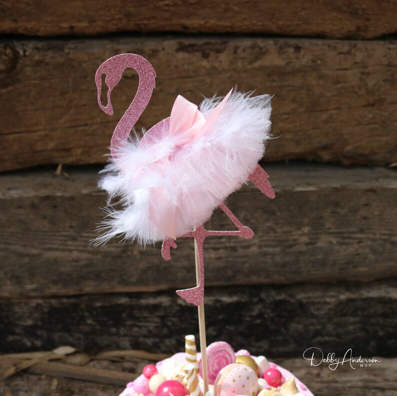 K&ouml;nnte beinhalten: Rosa glitzernder Flamingo-Kuchentopper mit einem wei&szlig;en T&uuml;ll-Tutu und einem rosa Band. Der Flamingo steht auf einem Bein und hat den Kopf zur Seite gedreht.