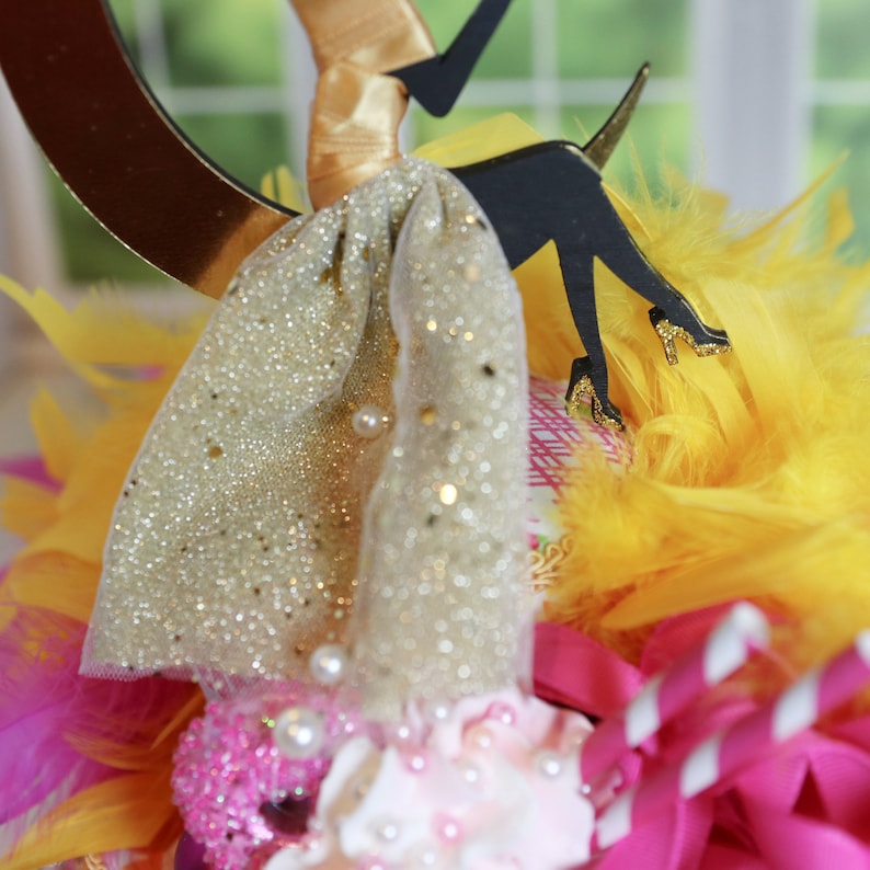 Peut inclure: Une d&eacute;coration de g&acirc;teau repr&eacute;sentant la silhouette noire d'une silhouette portant une jupe et des chaussures &agrave; paillettes dor&eacute;es, agr&eacute;ment&eacute;e de perles blanches, entour&eacute;e de plumes jaunes et roses, de rubans roses et de pailles ray&eacute;es.