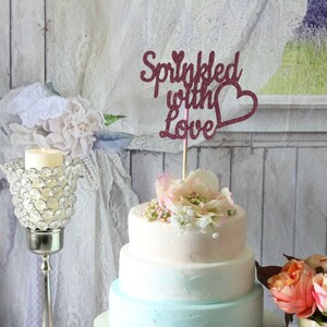 May include: A three-tiered wedding cake decorated with a pink and blue ombre frosting. The cake is topped with a pink glitter cake topper that says "Sprinkled with Love" with a heart.